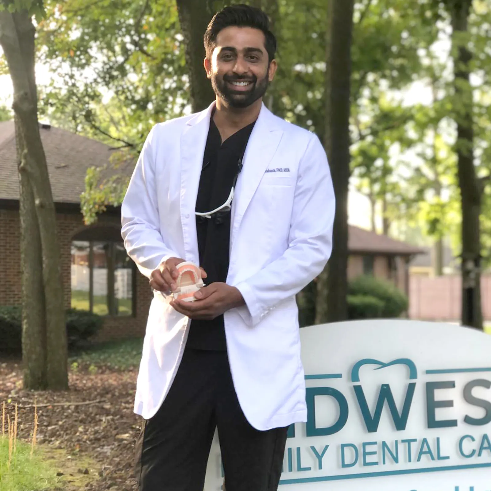 Dr. Varun Chintakunta, DMD, smiling in a white coat holding a dental model outside Midwest Family Dental Care sign, emphasizing his expertise in patient care and advanced dentistry.