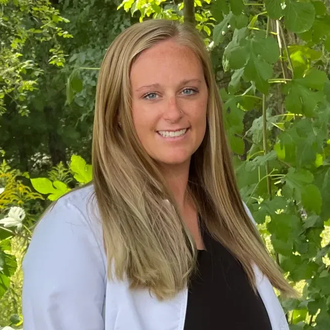 Dr. Janel Hackbardt smiling outdoors in a dental coat, surrounded by greenery, representing her commitment to patient care in Grand Rapids, Michigan.