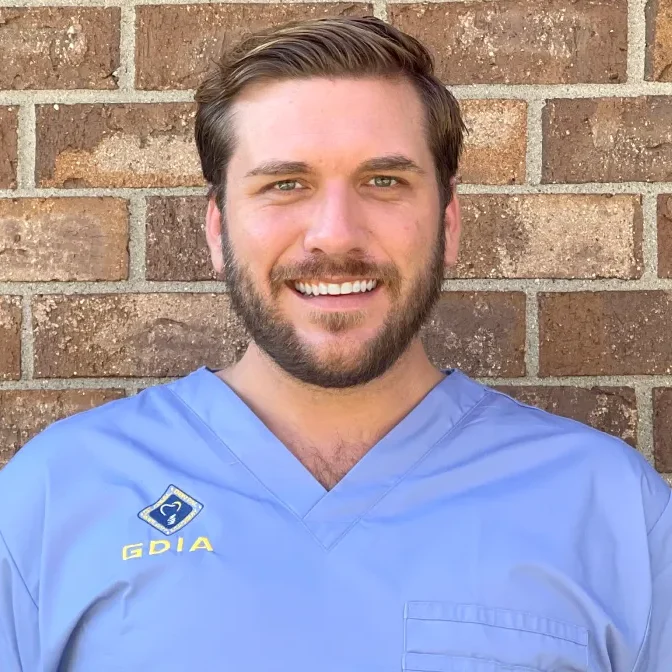 Dr. O'Donnell smiling in dental scrubs against a brick wall, representing compassionate dental care and commitment to patient wellness at Midwest Family Dental Care.