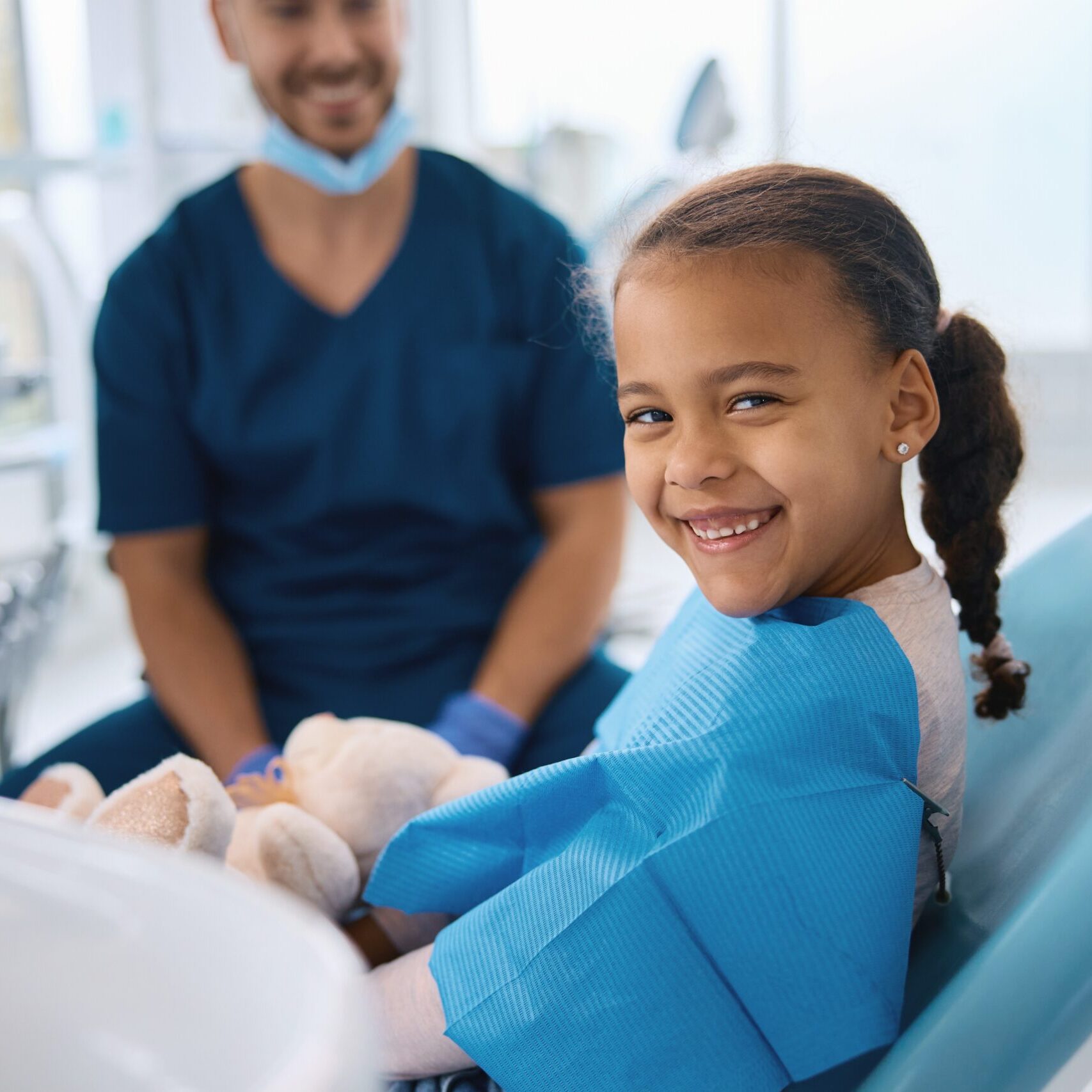 Smiling young girl in dental chair wearing blue bib, holding stuffed animal, with dentist in background, representing pediatric dentistry at Midwest Family Dental Care in Kokomo.